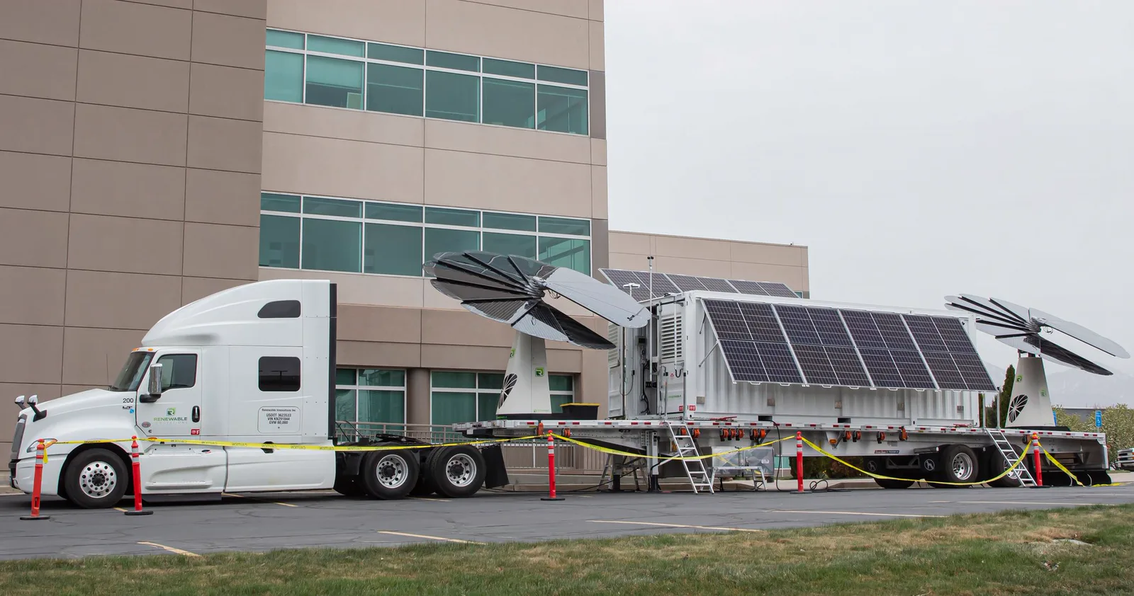 MEC Solar trailer parked beside an office building with its SmartFlower arrays deployed, hitched to a white semi tractor